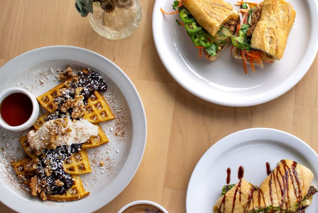 Wooden table topped with various plates including a french roll sandwich with cucumbers and jalapenos and waffles with whipped cream, sauce, and walnuts