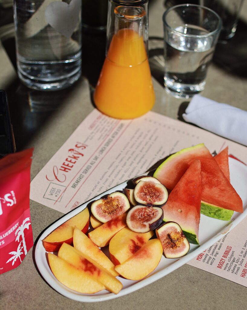 White plate with cut up figs, nectarines, and watermelon sitting on a table with a menu, a carafe of orange juice, and some glassware