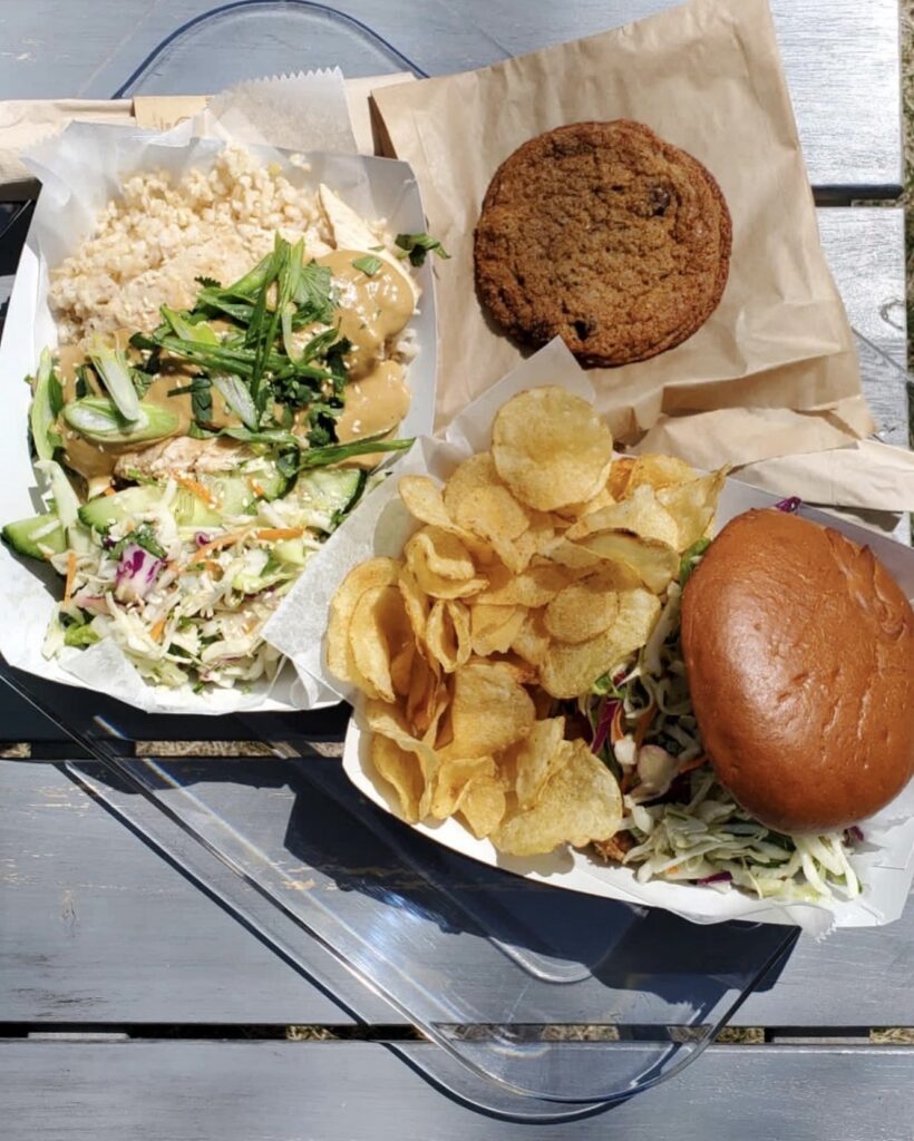 Overhead shot of a cookie, a rice bowl with scallions, cabbage, cucumbers,and chicken and a burger with a cabbage slaw and potato chips