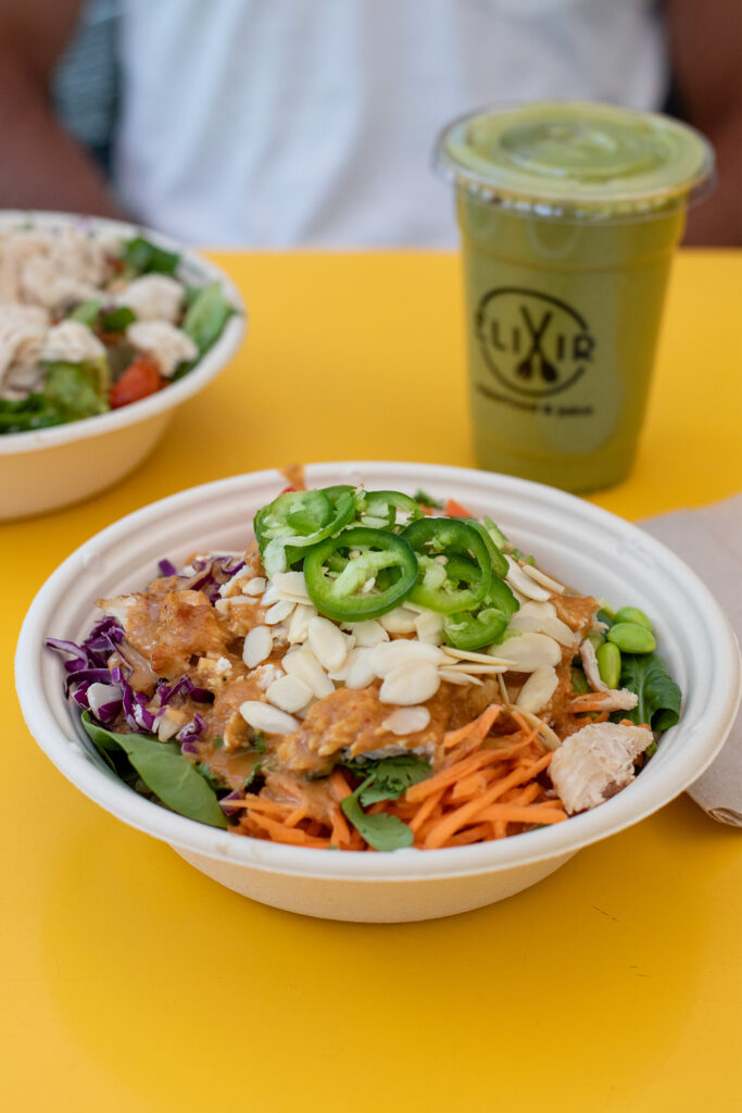 Yellow table with a container filled with carrots, chicken, jalapenos, spinach, cabbage, almonds, and sauce with a green smoothie in the background