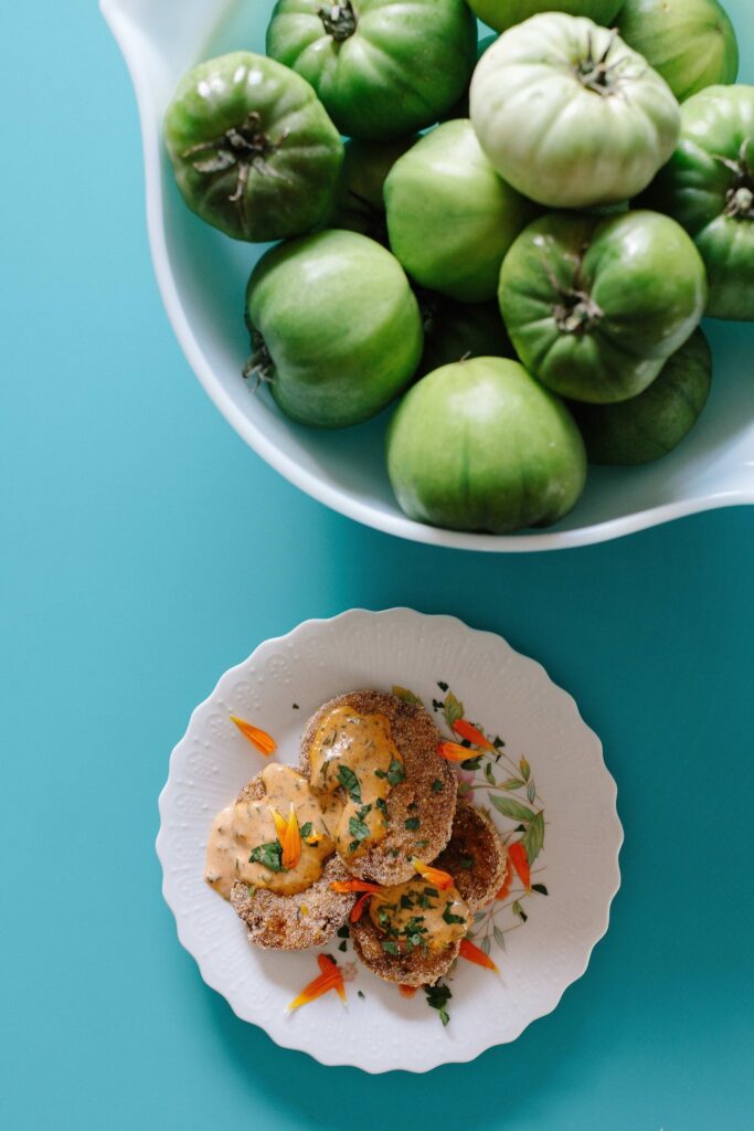 Blue table with a bowl of green tomatoes and a white dish with fried breaded tomatoes