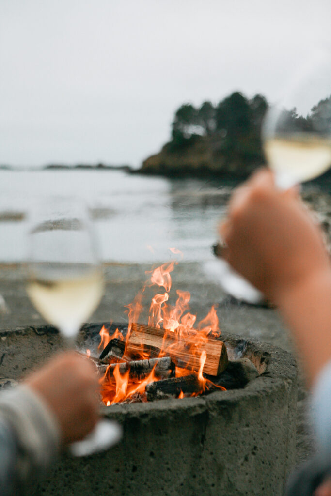 Two people with wine glasses around a fire at the beach with water in the background