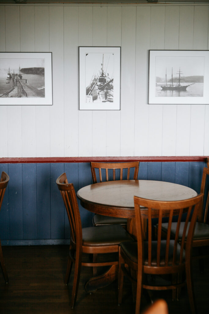 Wooden table with chairs in front of a blue and white wooden wall hung with black and white photos of a cafe at the Little River Inn