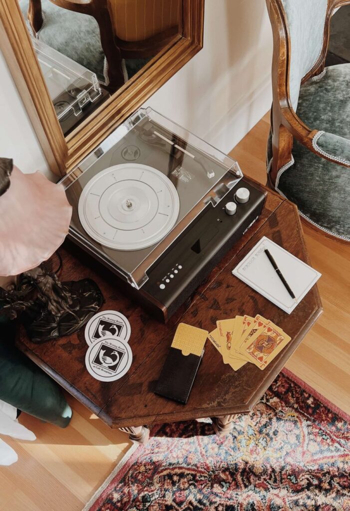 Wooden side table with vintage record player, lamp with pink lampshade, coasters, and playing cards Interior of the Nicholson House