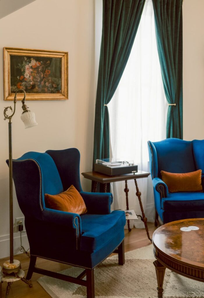 Blue velvet sitting chairs with copper throw pillows in a sitting room in front of a window with drapes at the Nicholson House