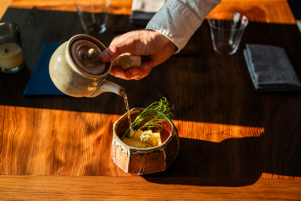 Hand pouring broth into a bowl of vegetables on a wooden table The Harbor House Inn, Restaurant, & Farm