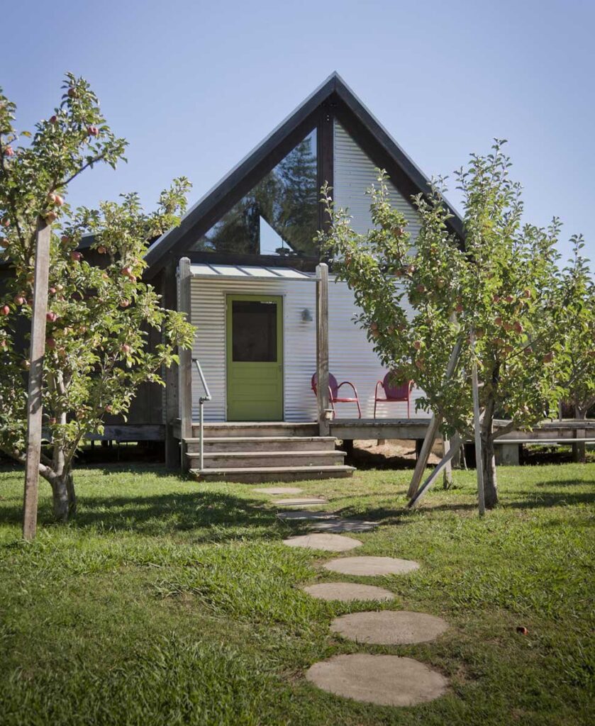 An a-frame corrugated metal and glass cabin with a green door and red sitting chairs in an apple grove 