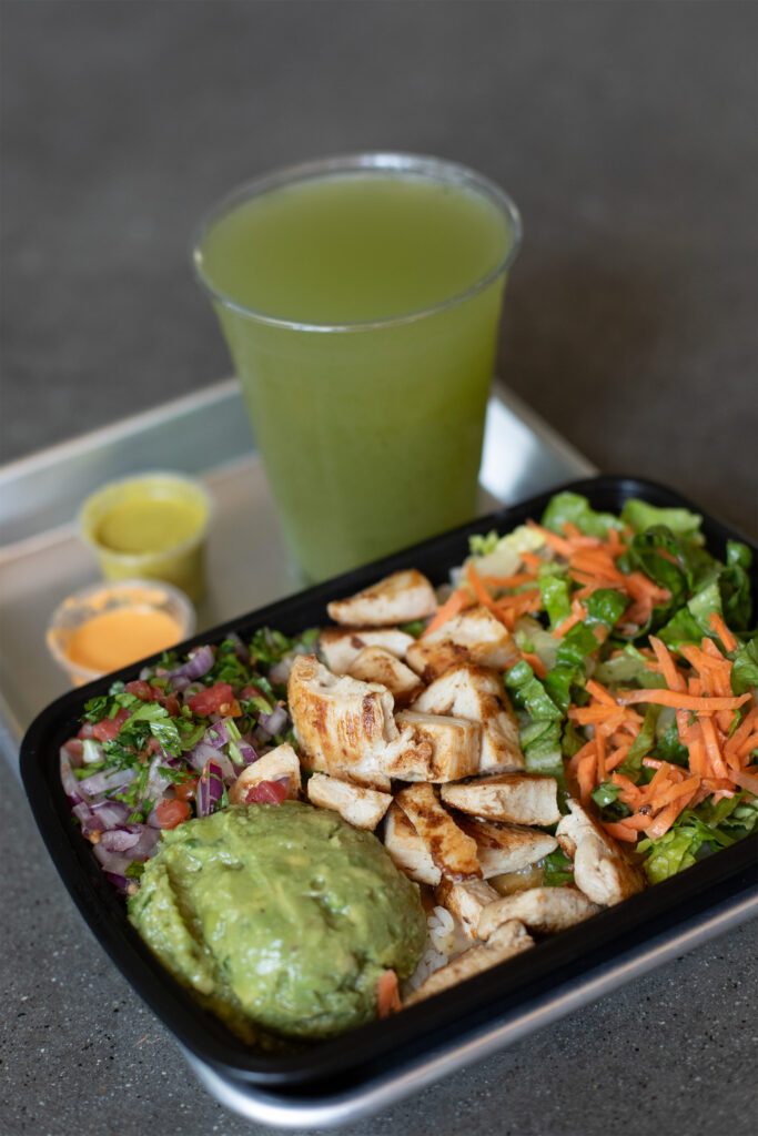 Shot of a black container with guacamole, chicken, pico de gallo salsa, greens, and carrots with sauces and a green drink in the background