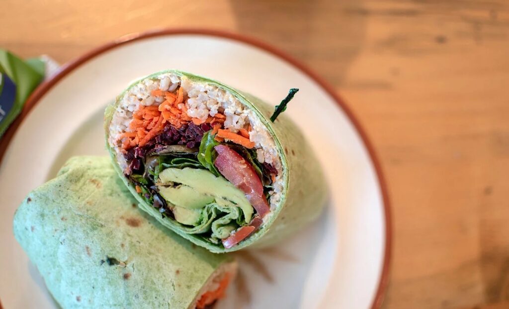 Wooden table with a white plate that has a halved wrap that's filled with rice, carrots, cabbage, tomatoes, greens, and avocado