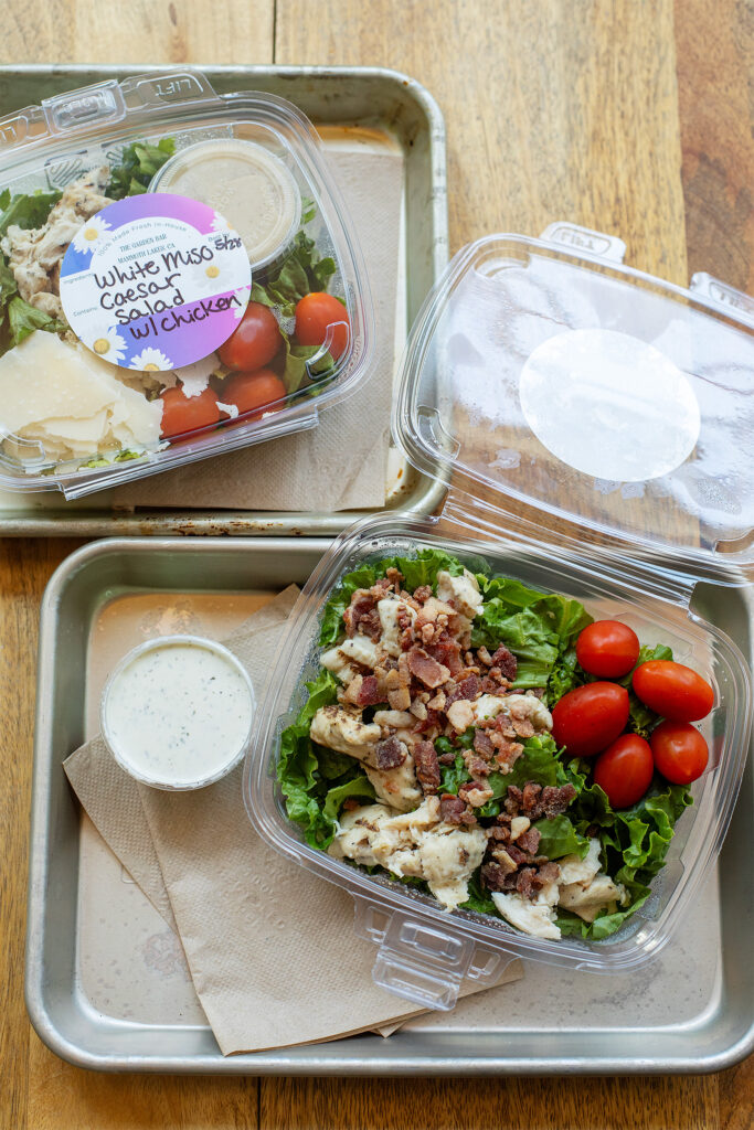 Wooden table with two containers of salad with greens, chicken, and tomatoes