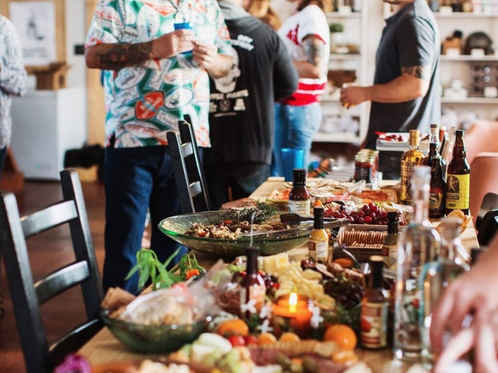 A table covered with food and drinks with people standing around in the background