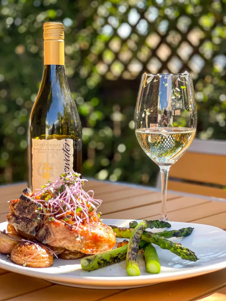 Wooden table with a plate with asparagus, potatoes, meat, and a bottle and glass of white wine in the background