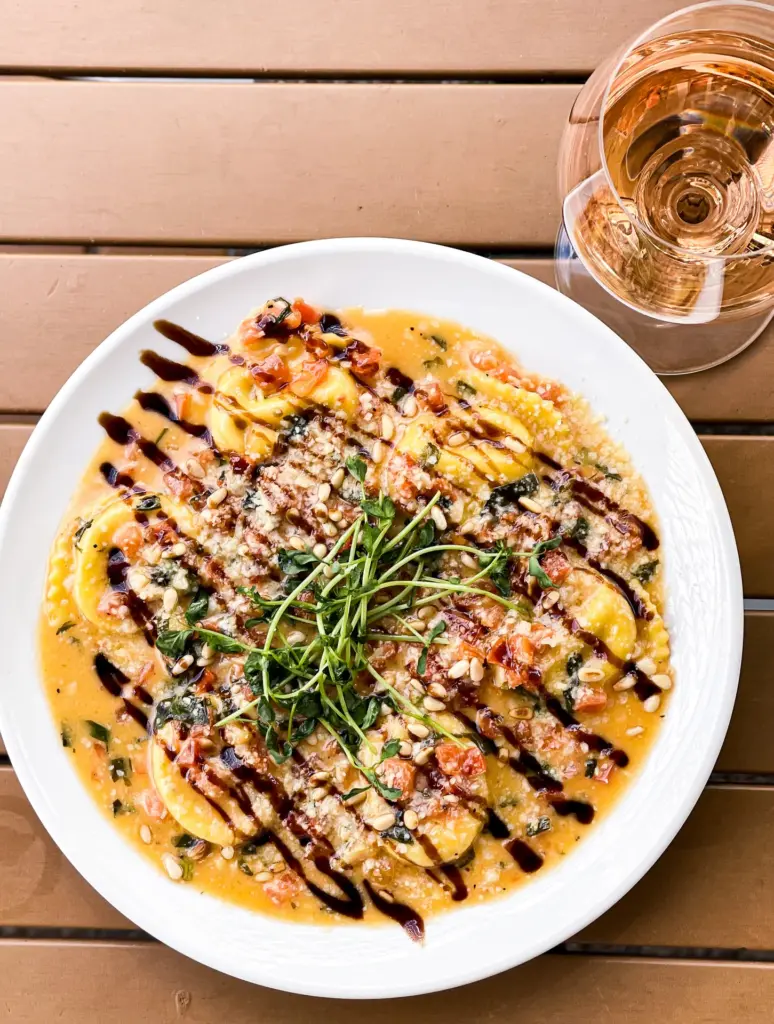 Overhead of a plate with ravioli pasta, yellow sauce, pine nuts, tomatoes, and herbs