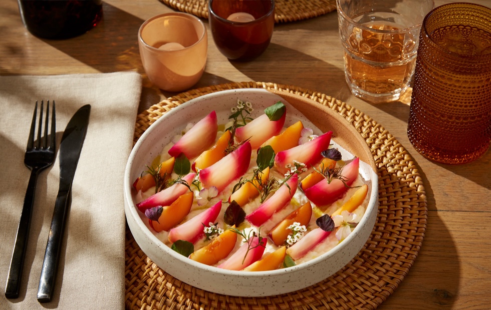 White bowl with various cut up plums on a wooden mat, a napkin, and glassware in the background