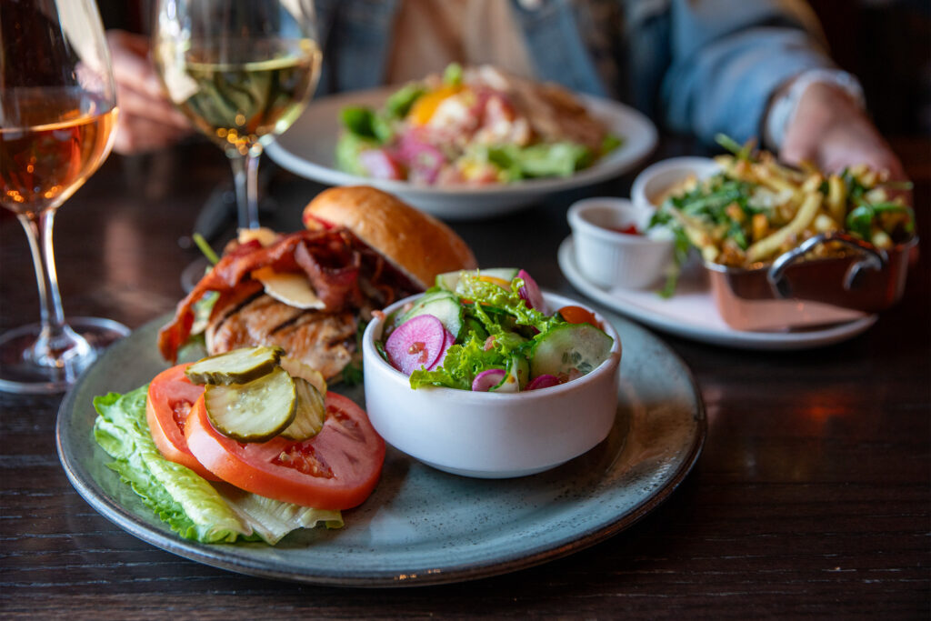 Chicken sandwich with a seasonal side salad, a side of fries with arugula, a salad, and glasses of wine