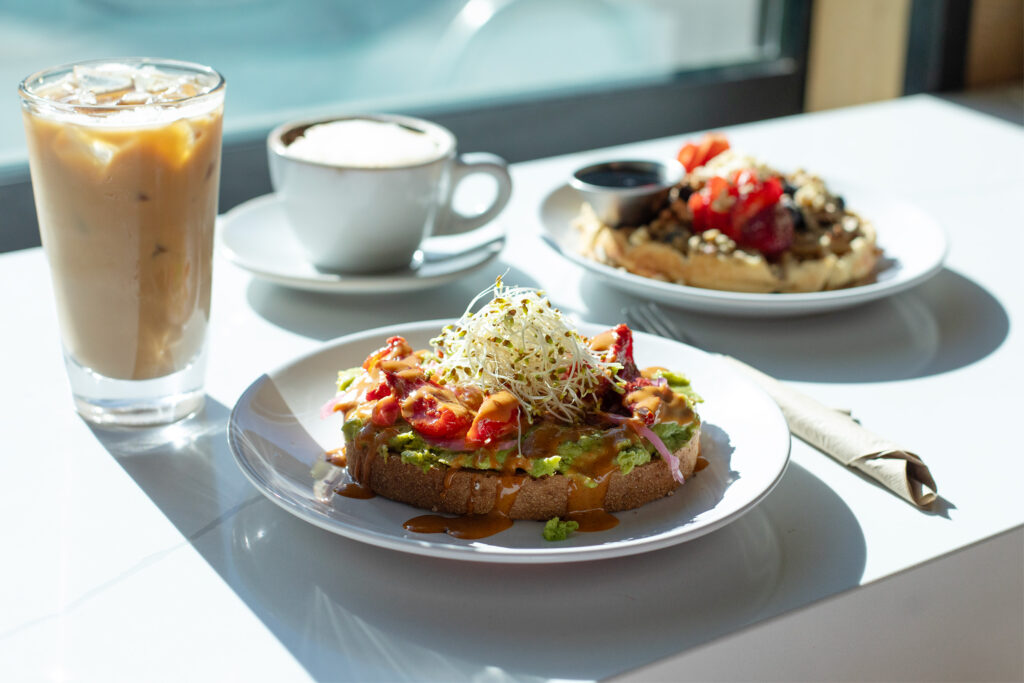 Avocado toast with tomatoes and sprouts on a table with coffees and a waffle with berries