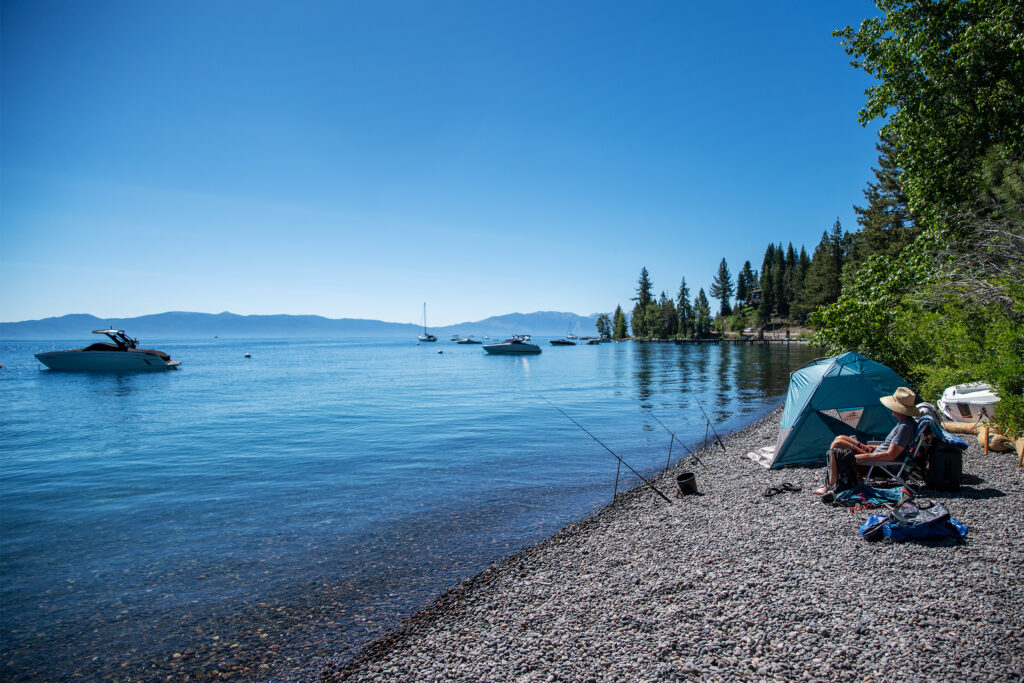 Person sitting on a lakeshore next to a tent with fishing rods in the water