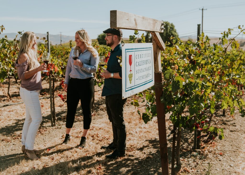 Group of people talking and holding wine glasses standing next to a sign while in a vineyard in Livermore wine country