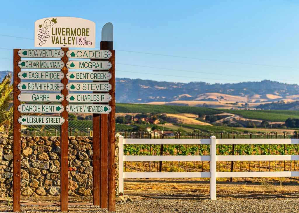 Winery directional signs next to a white fence and with vineyards and hills in the background in Livermore wine country